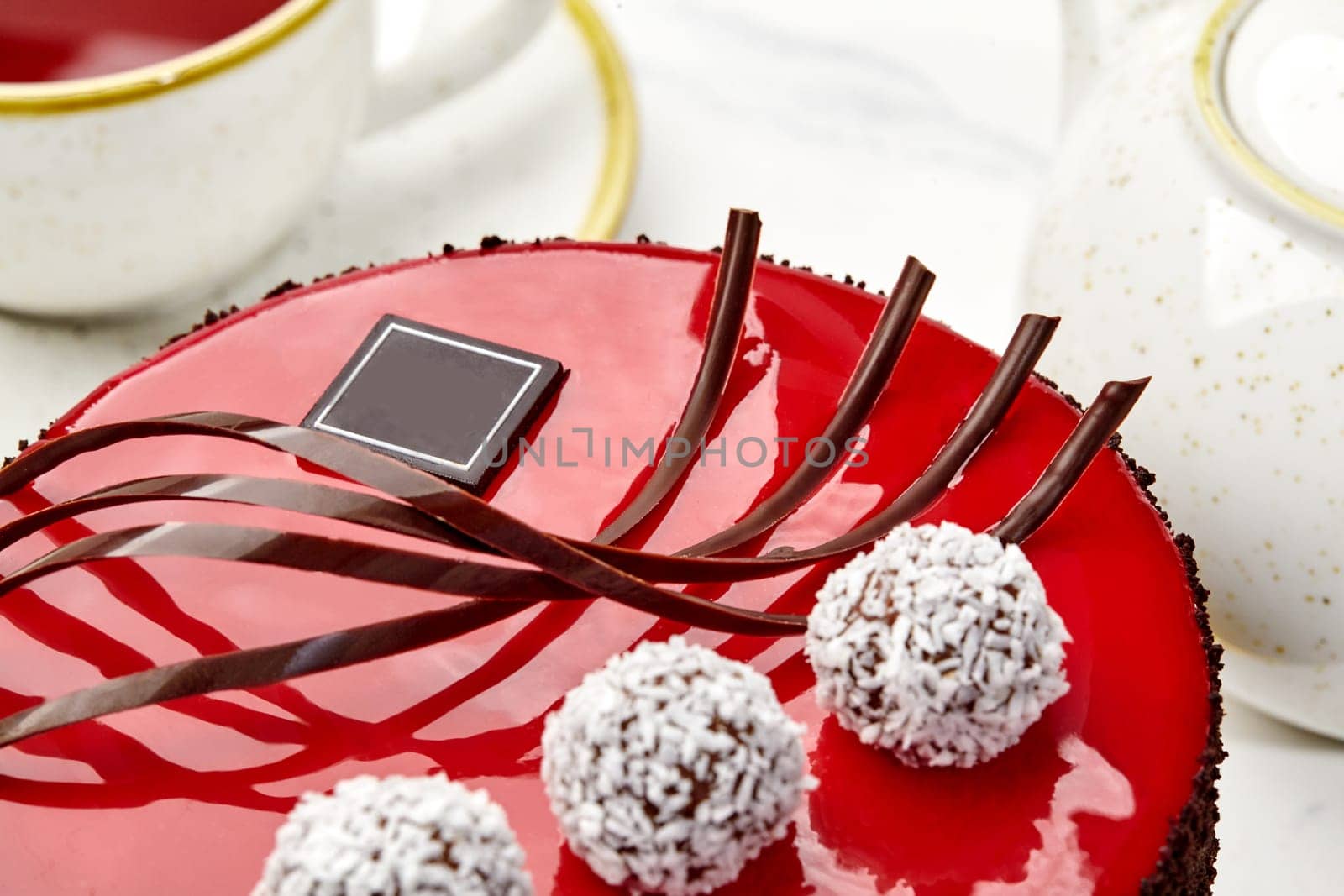 Closeup of artfully decorated cake with red mirror glaze, glossy dark chocolate swirls and white coconut truffles served with tea for special occasion