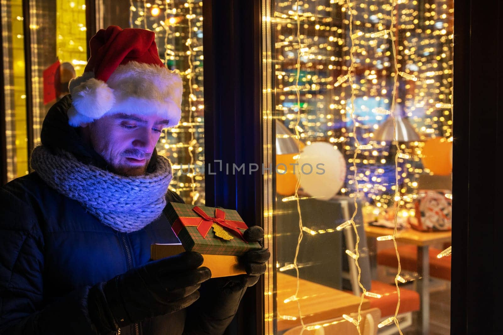 Man in Santas hat with gifts box near illuminated cafe window.