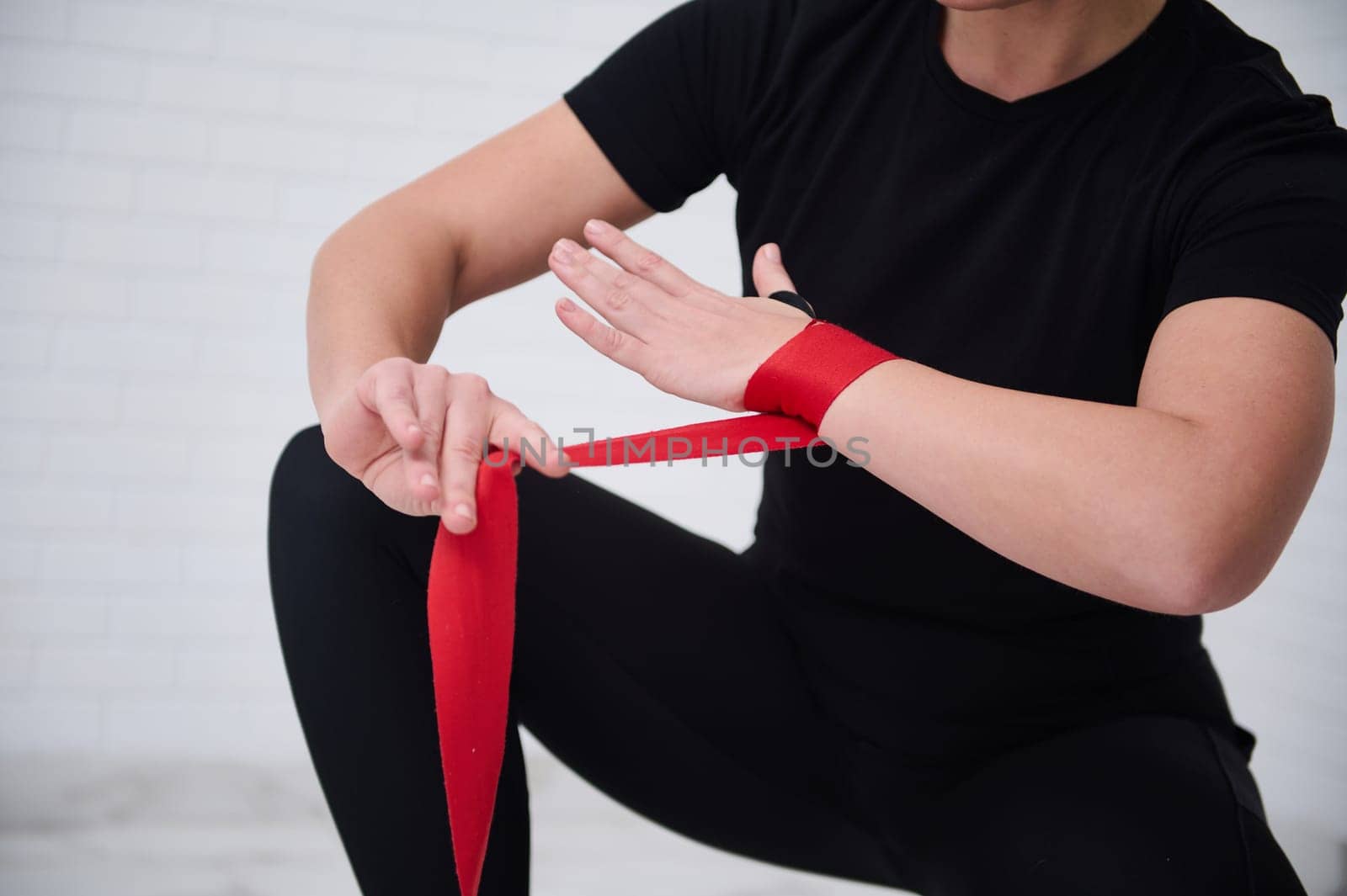Close-up strong woman boxer fighter preparing boxing bandages, wrapping her wrist and hands with a red tape, before putting on boxing gloves, getting ready for training, isolated over white background