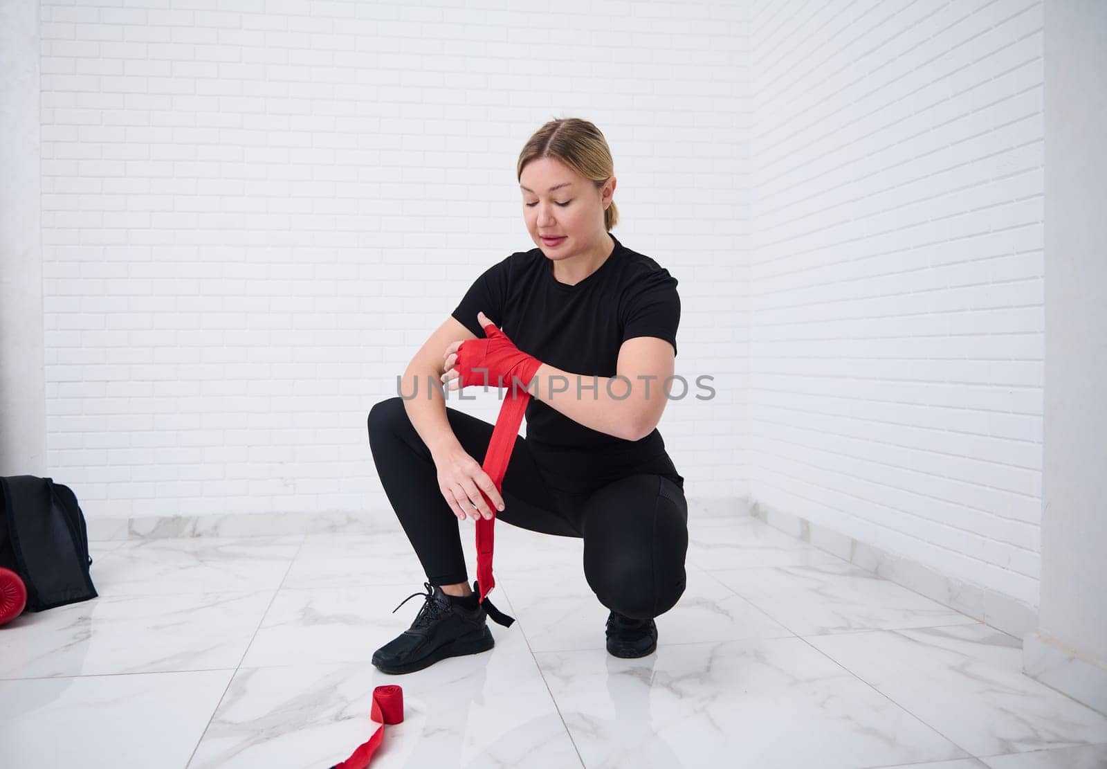 Full size shot of Caucasian confident female fighter in black sports wear, tying tape around her hand before get boxing gloves preparing to boxing practice. Martial art and combat concept. Top view