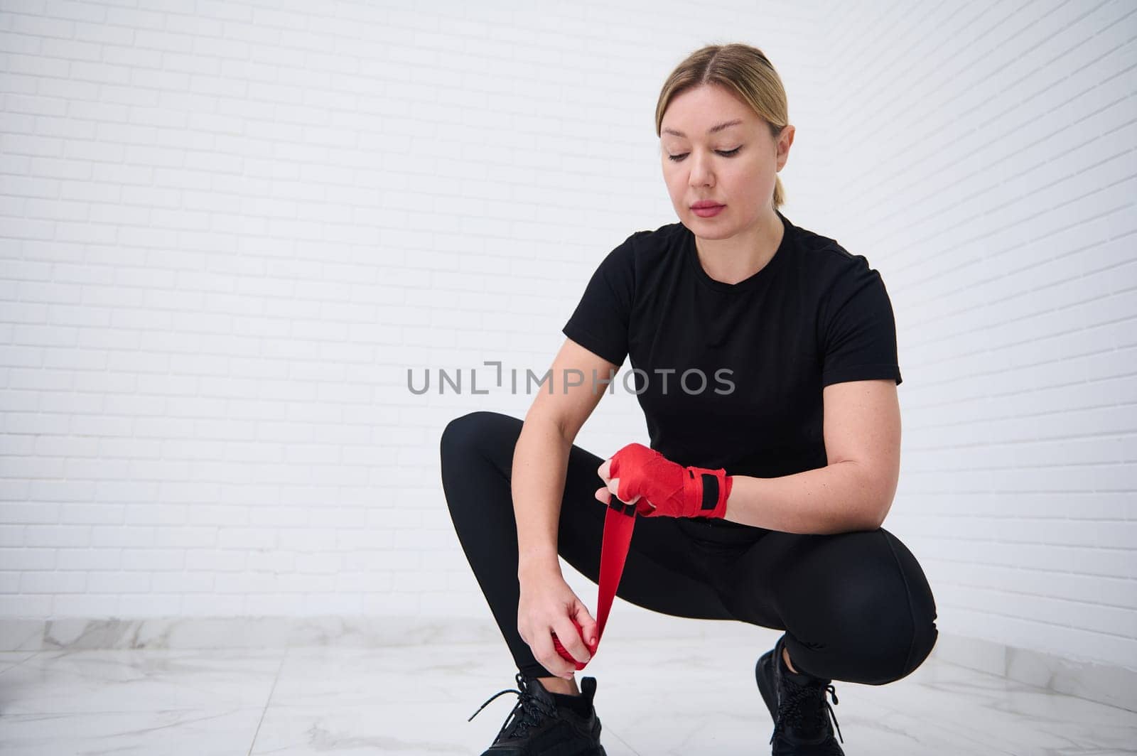 Caucasian young woman boxer fighter preparing boxing bandages, wrapping her wrist and hands with a red tape, before putting on boxing gloves, getting ready for training, isolated over white background