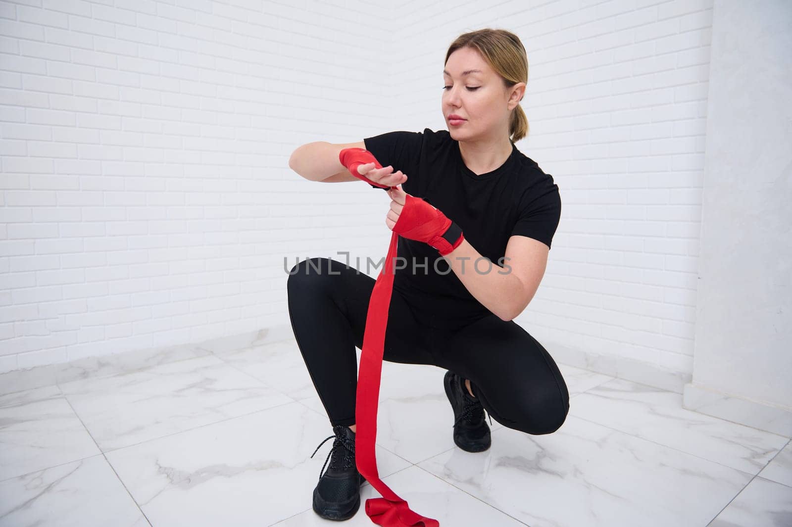 Authentic portrait of determined confident European young woman fighter 40s, in sports clothes, sitting in squat position, typing red tapes over white background, preparing for boxing training