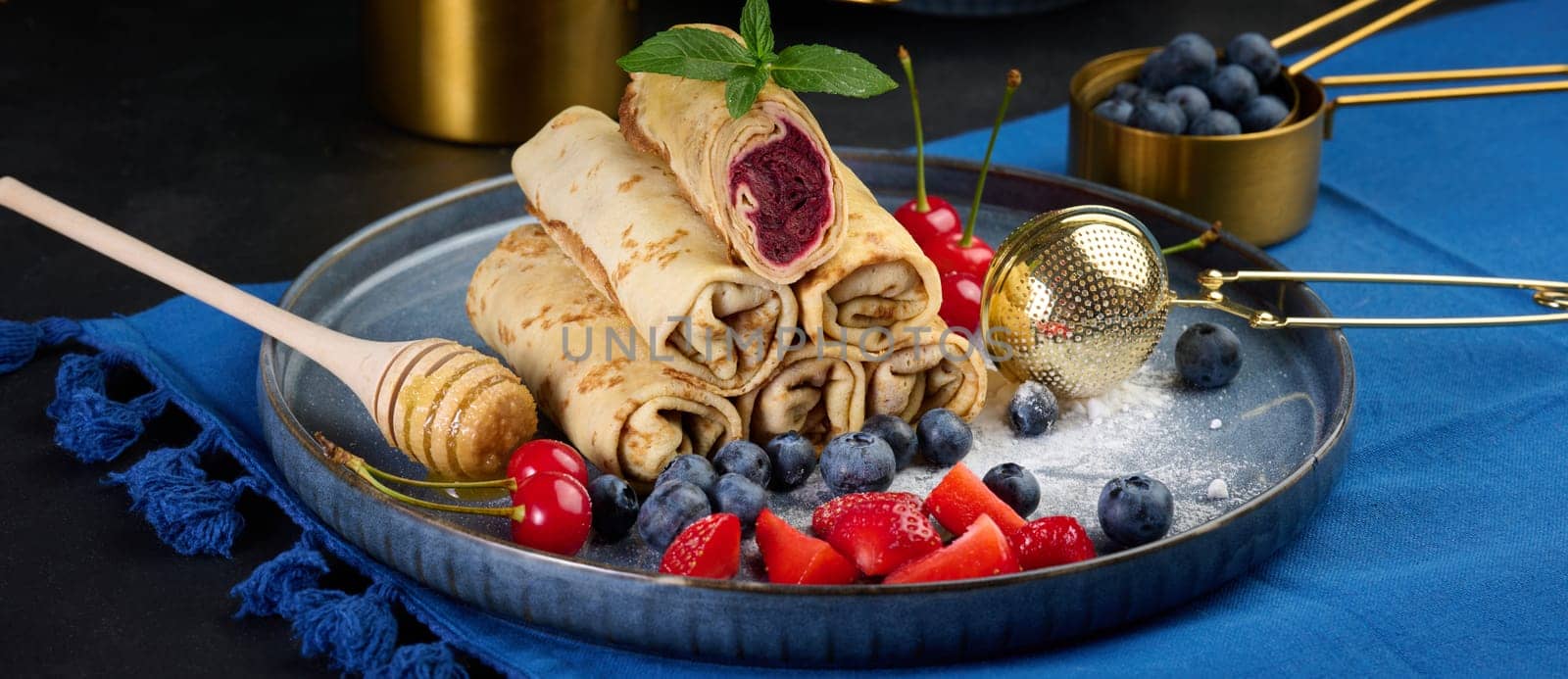 Crepes stacked in a pile, one of which is cut and filled with berry filling. The plate is also decorated with blueberries, strawberries, and cherries, a honey dipper with honey, a sprig of mint, and a golden sieve. A dark blue tablecloth.