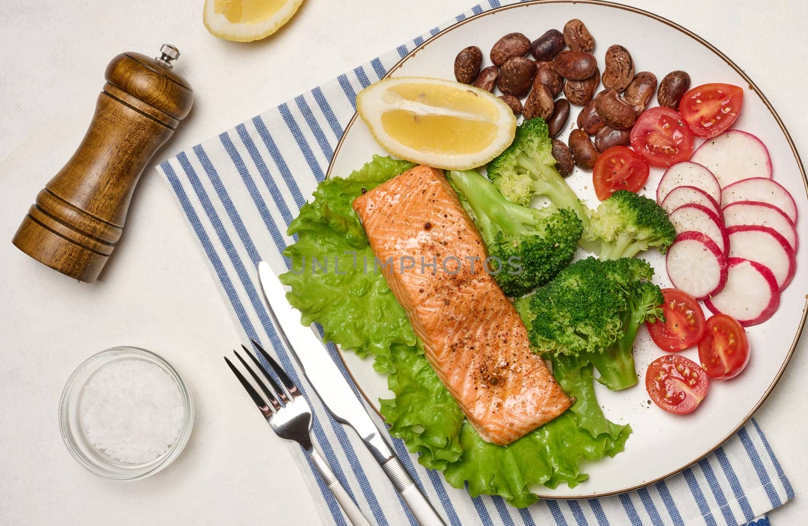 Healthy lunch with grilled salmon on green lettuce, next to vegetables, tomatoes, radishes, broccoli and a portion of beans. Accessories include a spice bowl, a bottle of olive oil, a fork and a pepper grinder piece.