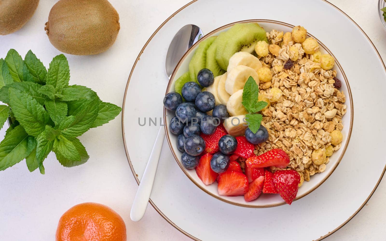 Granola with strawberries, kiwi, banana and blueberries in a round plate on a white table. Healthy and tasty food, top view