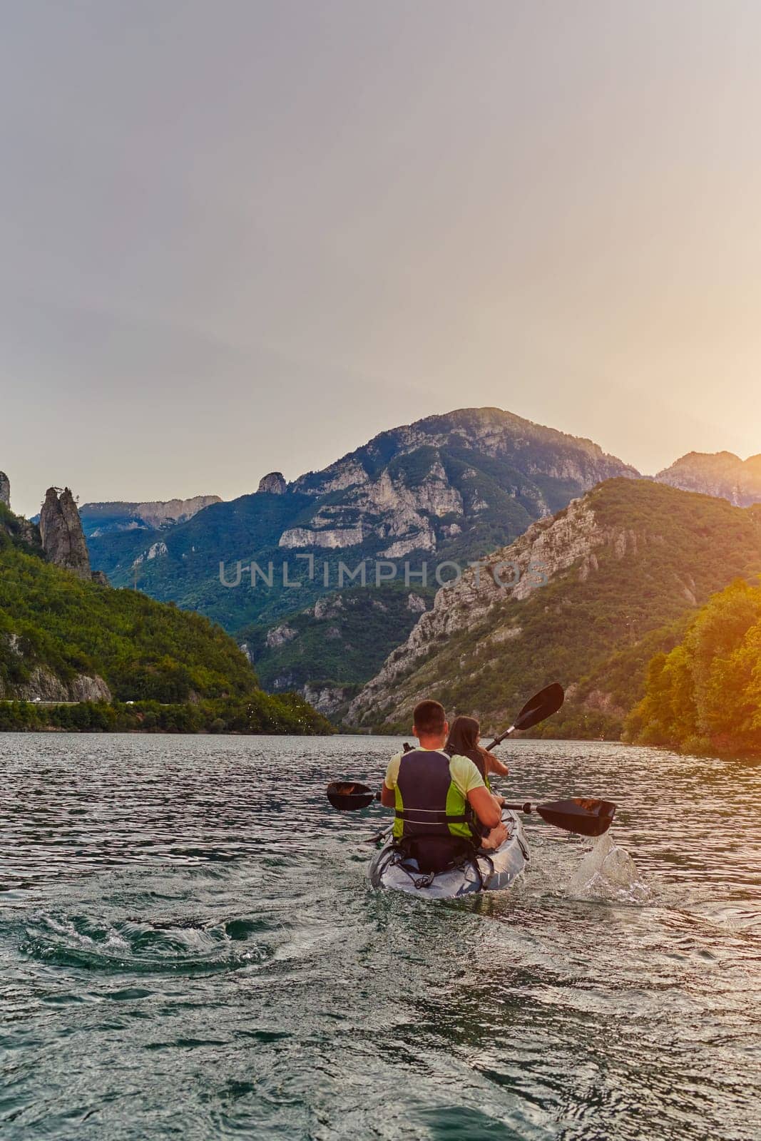 A young couple enjoying an idyllic kayak ride in the middle of a beautiful river surrounded by forest greenery in sunset time.