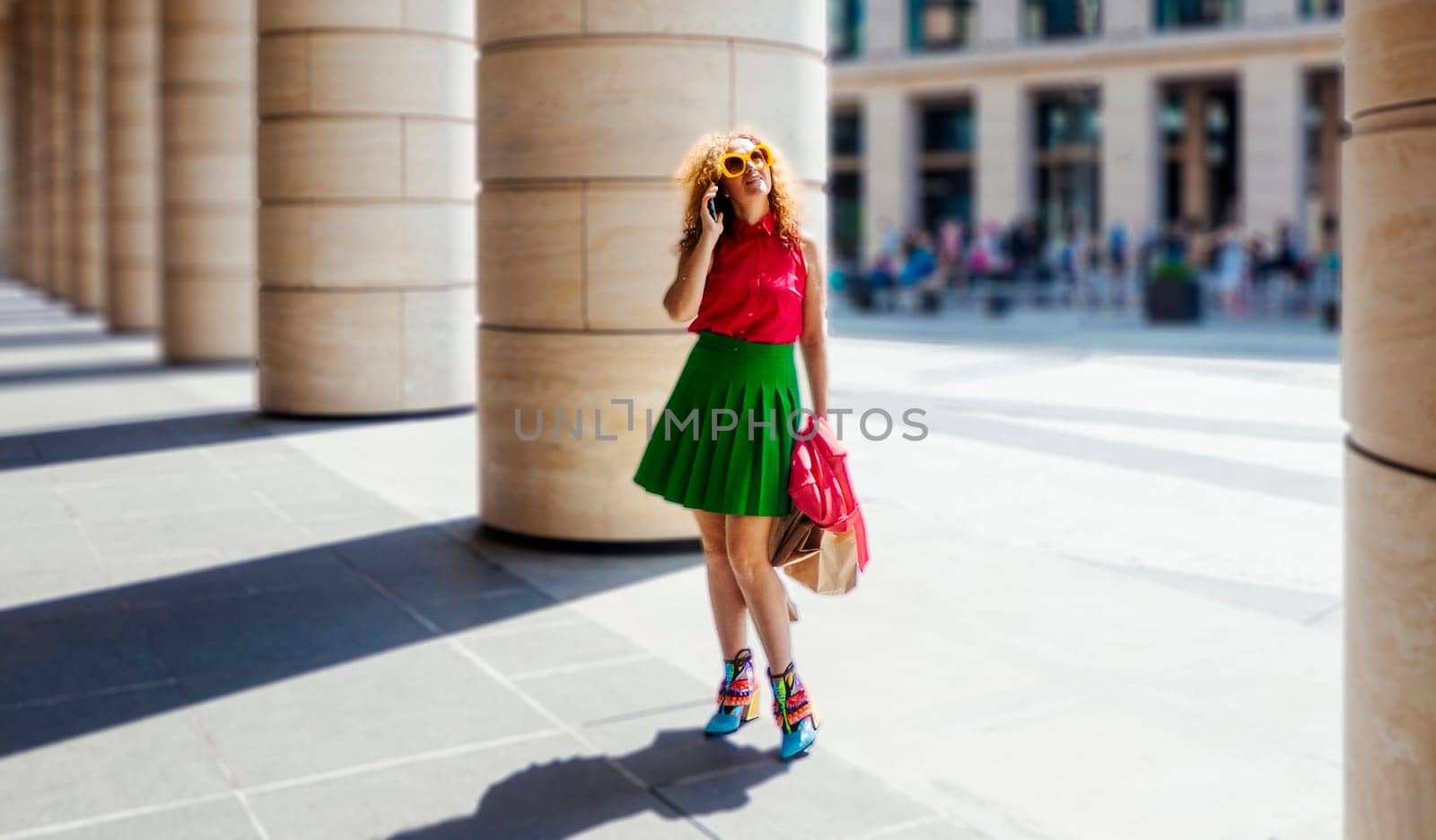 Content fashion female with bag walking on pavement near stone columns in sunlight and talk cell phone. the concept of freedom of tourism and fashion business