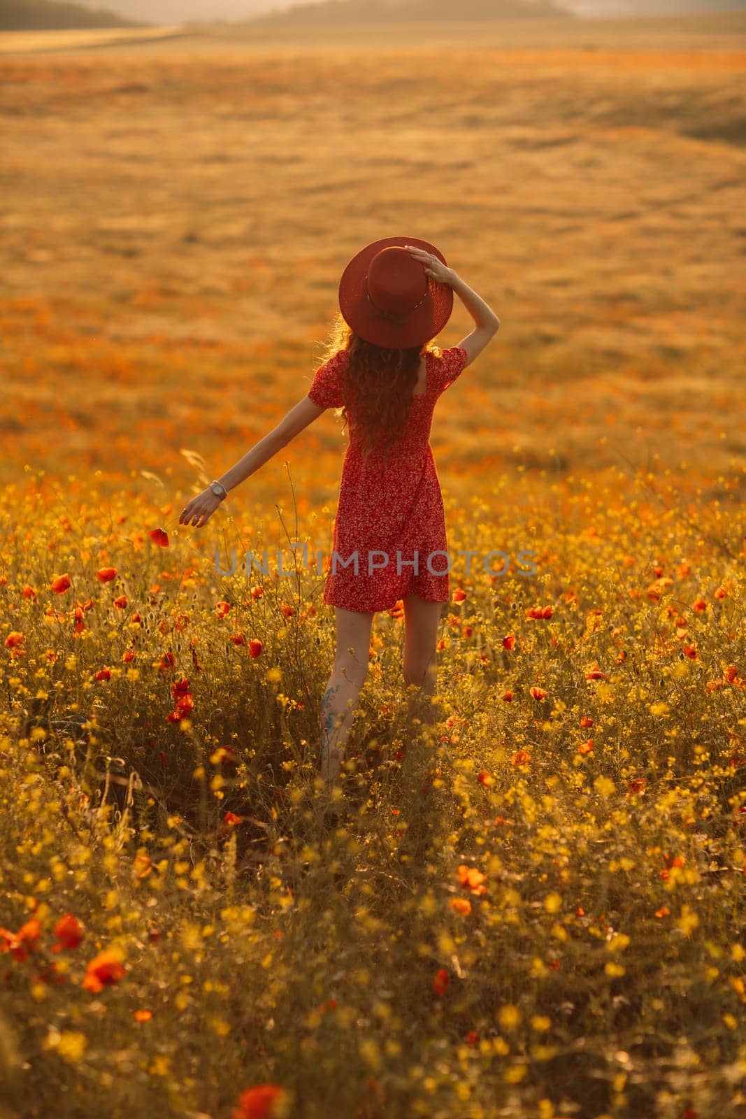 A woman in a red dress stands in a field of yellow flowers. She is wearing a red hat and has her hand up in the air. The scene is peaceful and serene, with the woman enjoying the beauty of the flowers