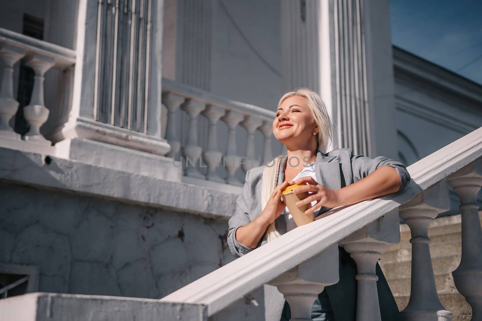 A woman is standing on a white railing with a cup of coffee in her hand. She is wearing a gray jacket and blue jeans. Concept of relaxation and leisure
