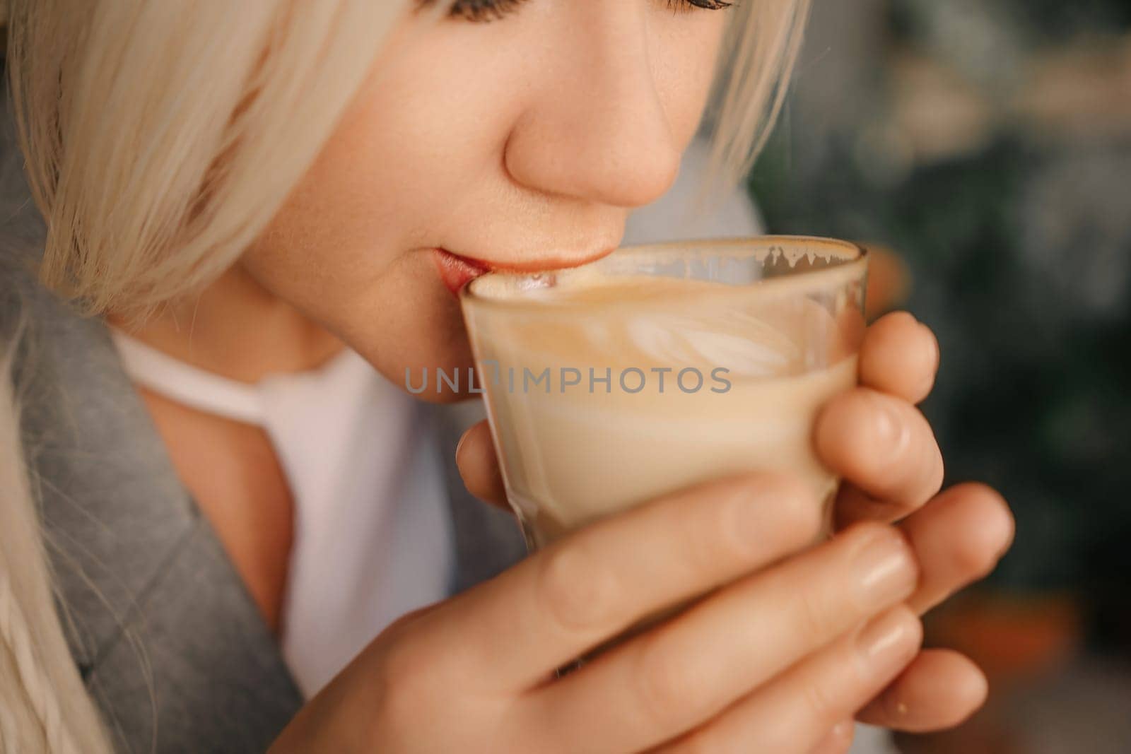 A blonde woman is drinking coffee from a glass. She is smiling and she is enjoying her drink