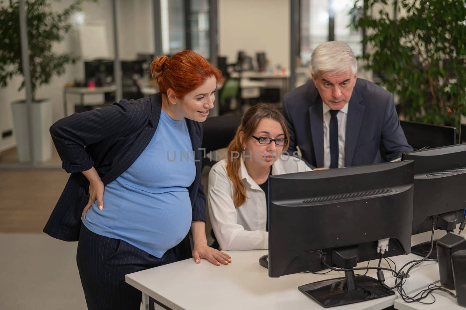 An elderly man, a Caucasian woman and a pregnant woman are discussing work issues at the computer