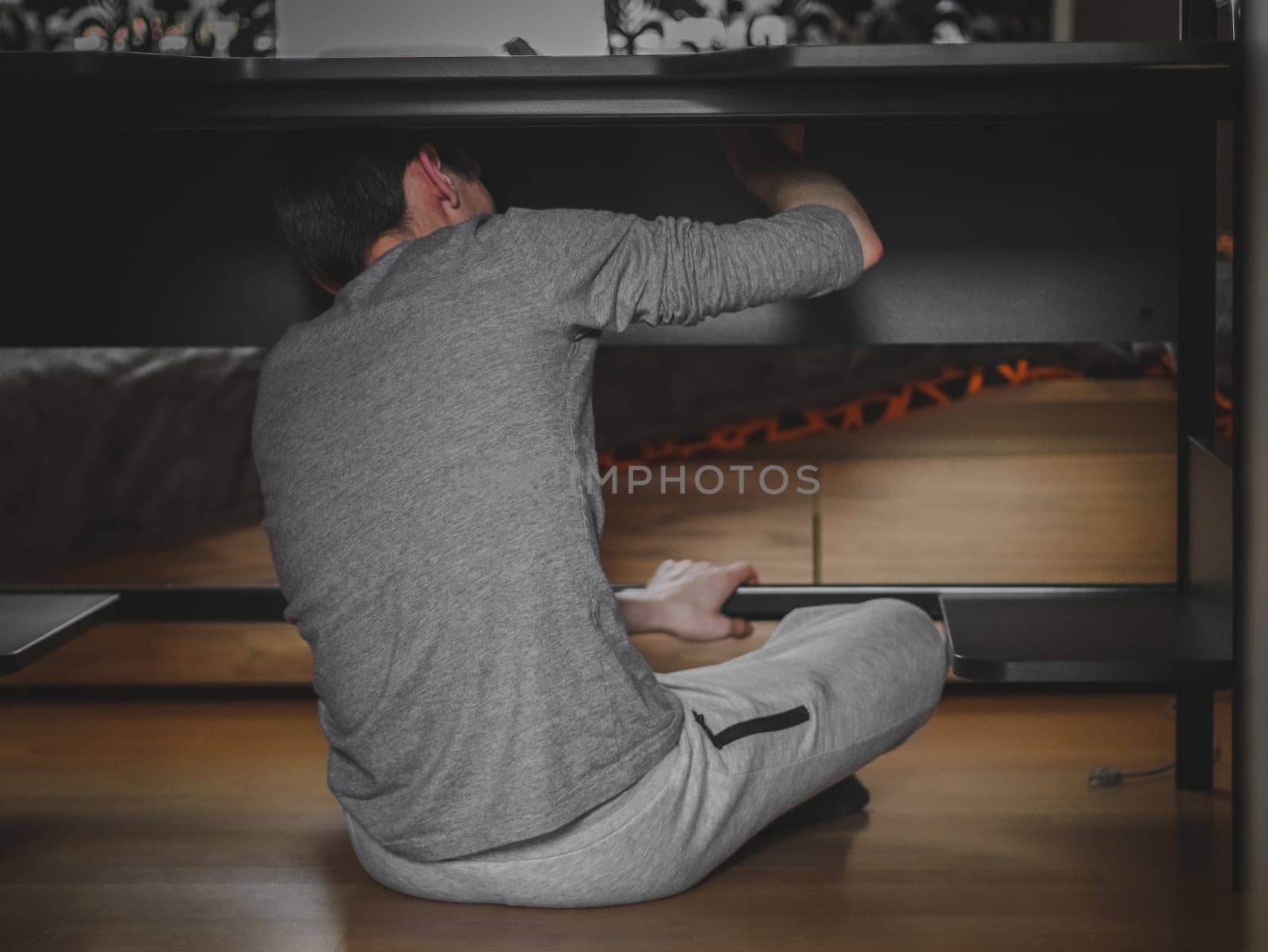 One Caucasian young man in gray clothes sits from his back on the floor under a black computer desk, manually tightening bolts during the day in the room, close-up side view with depth of field. Furniture assembly concept, assembly services.