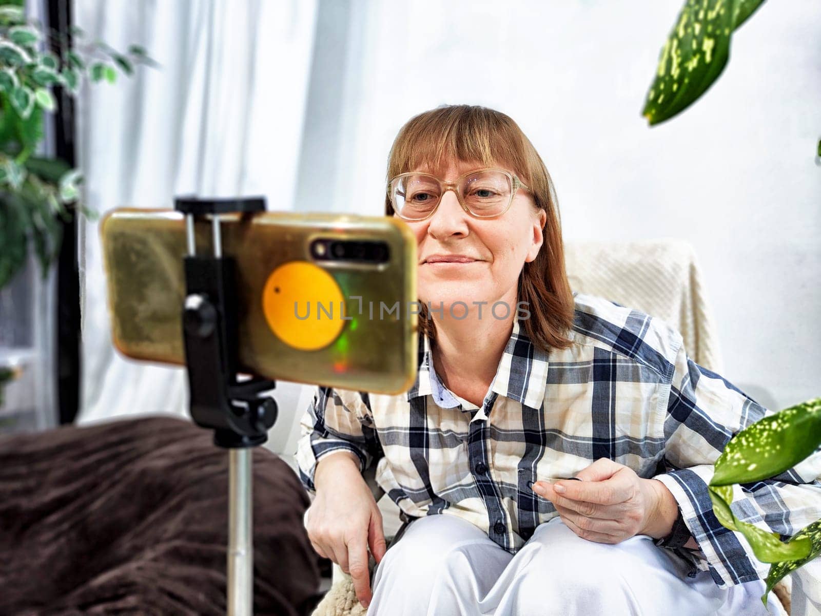 Smiling woman in glasses using a smartphone on a tripod for a video chat. A middle-aged woman posing and taking a selfie