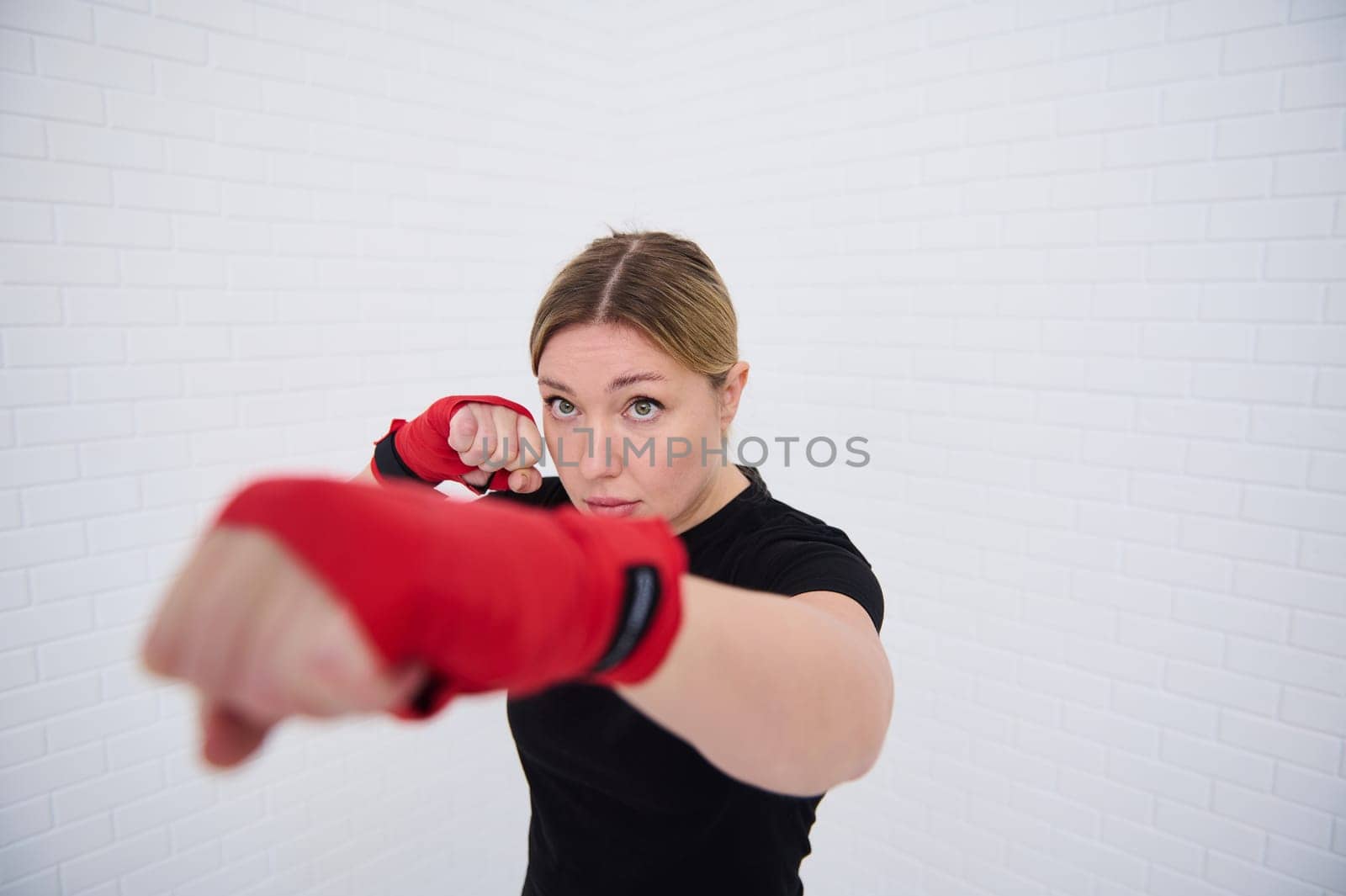 Confident European blonde woman dressed in black sportswear and red boxing gloves, punching forward while practicing box, isolated over white background. The concept of martial art and self-confidence