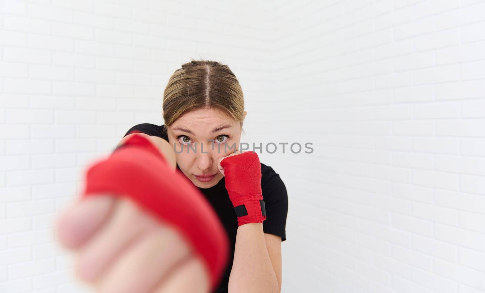 European blonde confident determined female boxer wearing boxing gloves, practicing punching forward the camera, isolated on white background. Blurred fists of hands on the foreground. Copy ad space.
