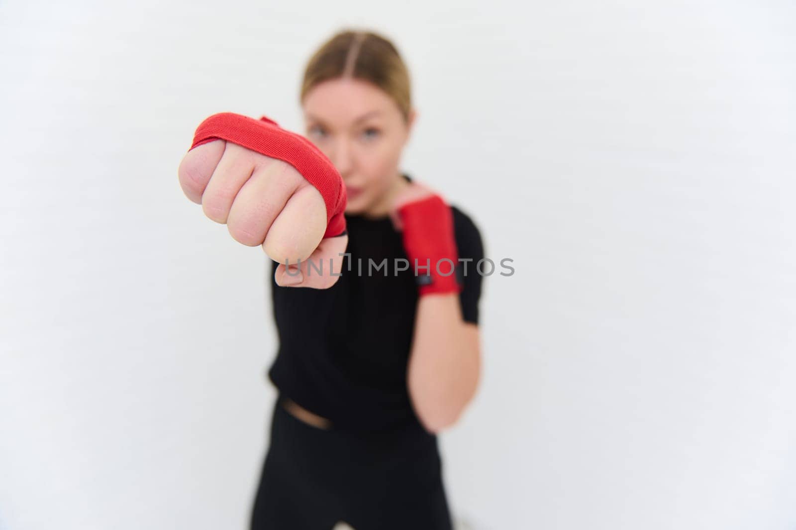 Selective focus on the fist of of a blurred young female boxer fighter in red boxing gloves, punching forward the camera, isolated over white background. Boxing training. Martial art. People and sport