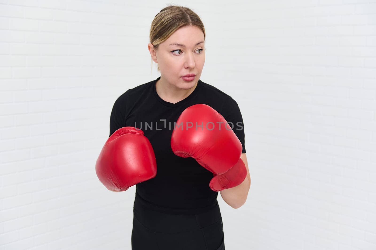 Authentic portrait of European female boxer with red boxing gloves, looking aside, isolated over white background with copy space. Pretty woman exercising during box workout indoors