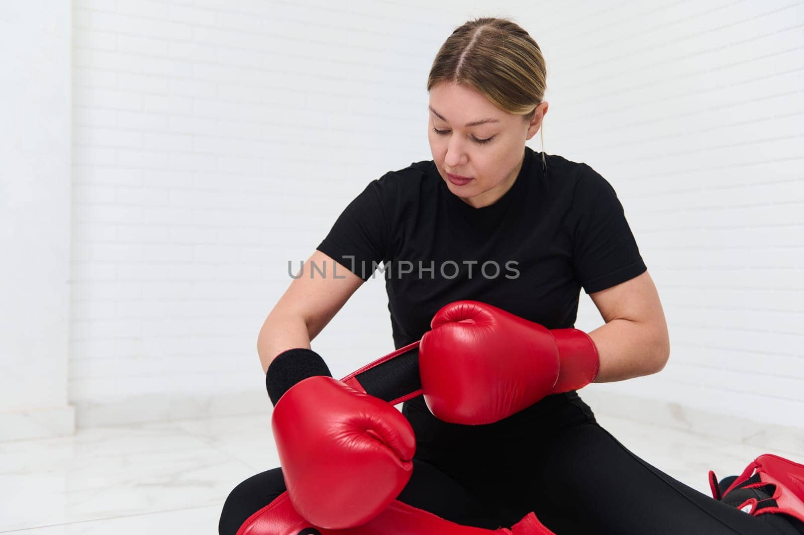 Determined European woman boxer, fighter in black sports wear, putting on boxing equipment and gloves, sitting on the floor, isolated over white wall background. Martial art, people and sport concept
