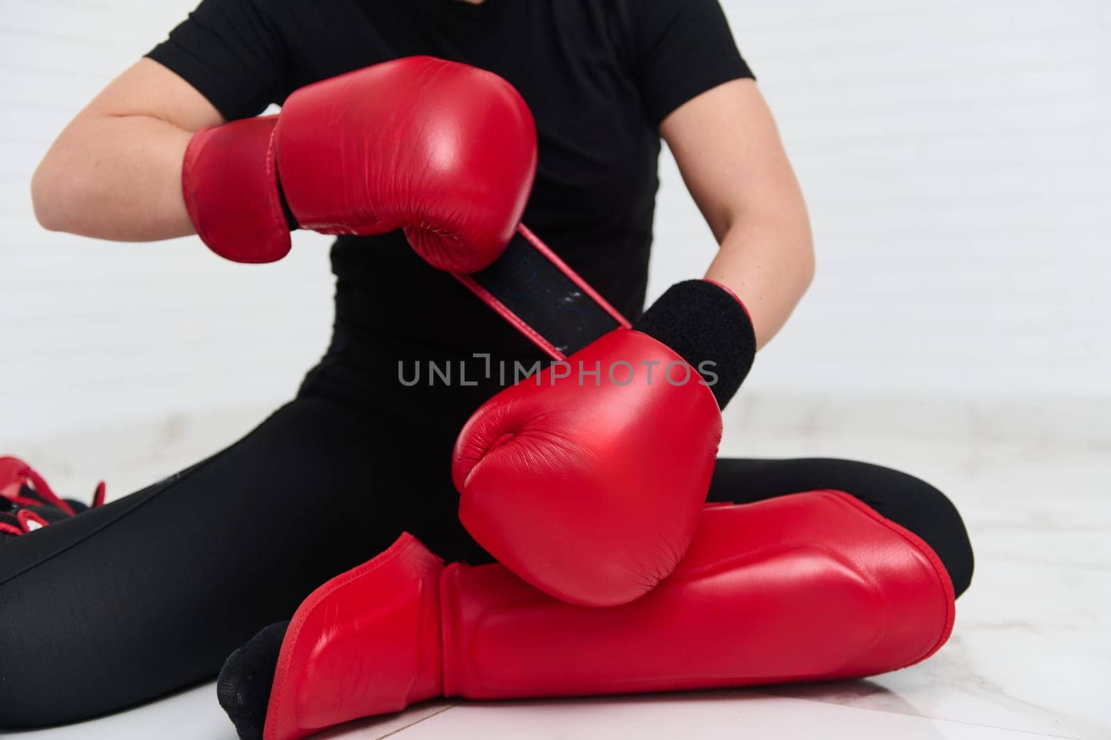 Cropped view of female boxer, fighter in black sports wear, putting on boxing equipment and gloves, sitting on the floor, isolated over white wall background. Active people and sport concept