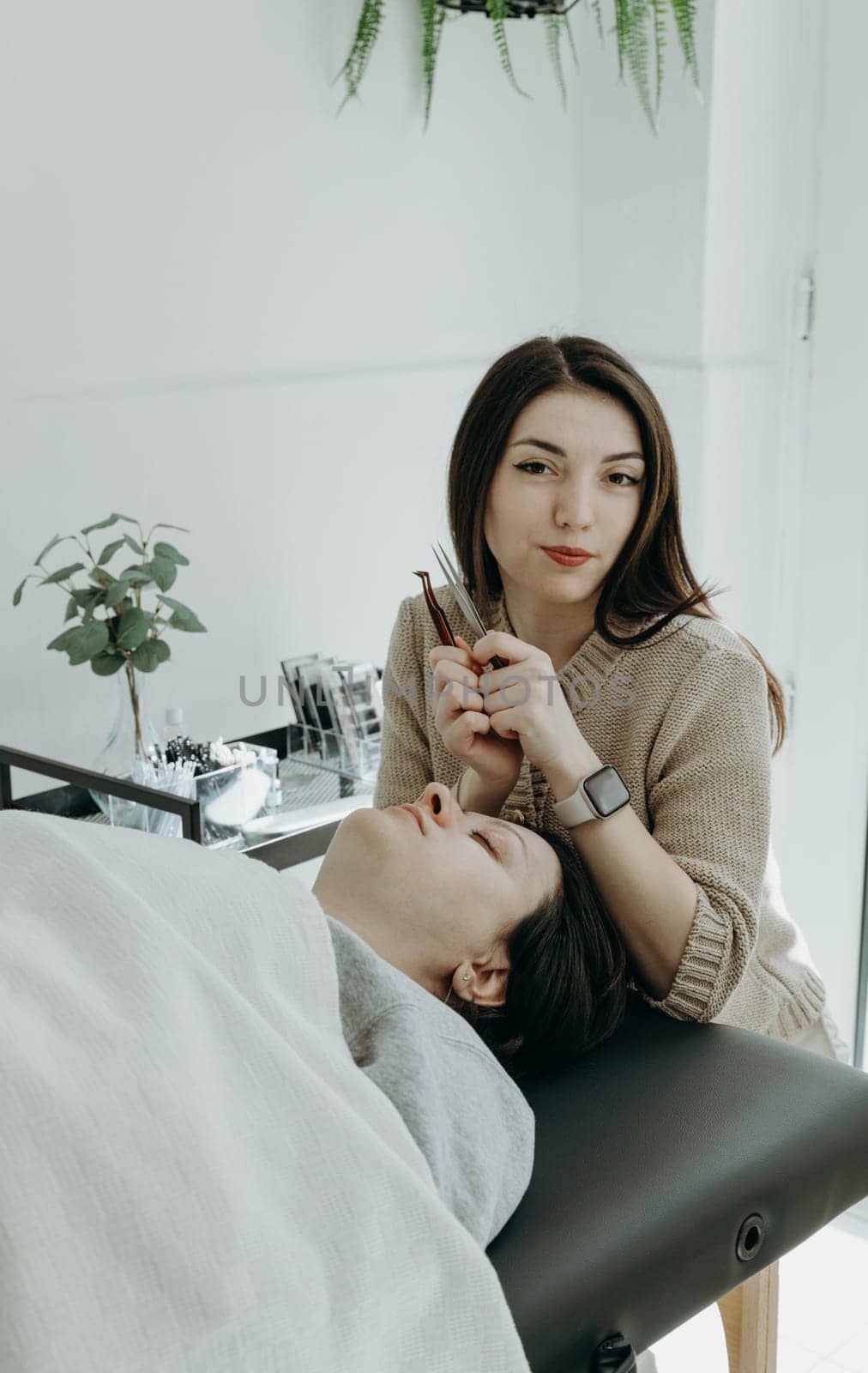 Portrait of one young beautiful Caucasian cosmetologist girl with a smile holding tools for eyelash extensions in her hands and a female client lying on the cosmetology table in the office on a spring day, side view close-up. Concept of eyelash extensions, beauty industry.