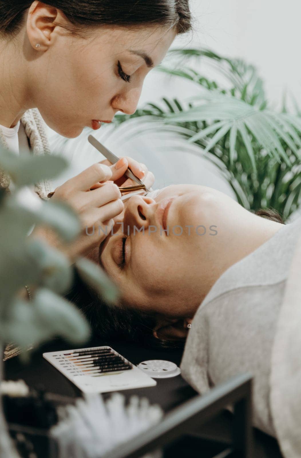 Portrait of one young beautiful Caucasian brunette girl, a cosmetologist glues artificial eyelashes with tweezers to the left of a female client lying on a cosmetology table in the office on a spring day, side view close-up. Concept of eyelash extensions, beauty industry.