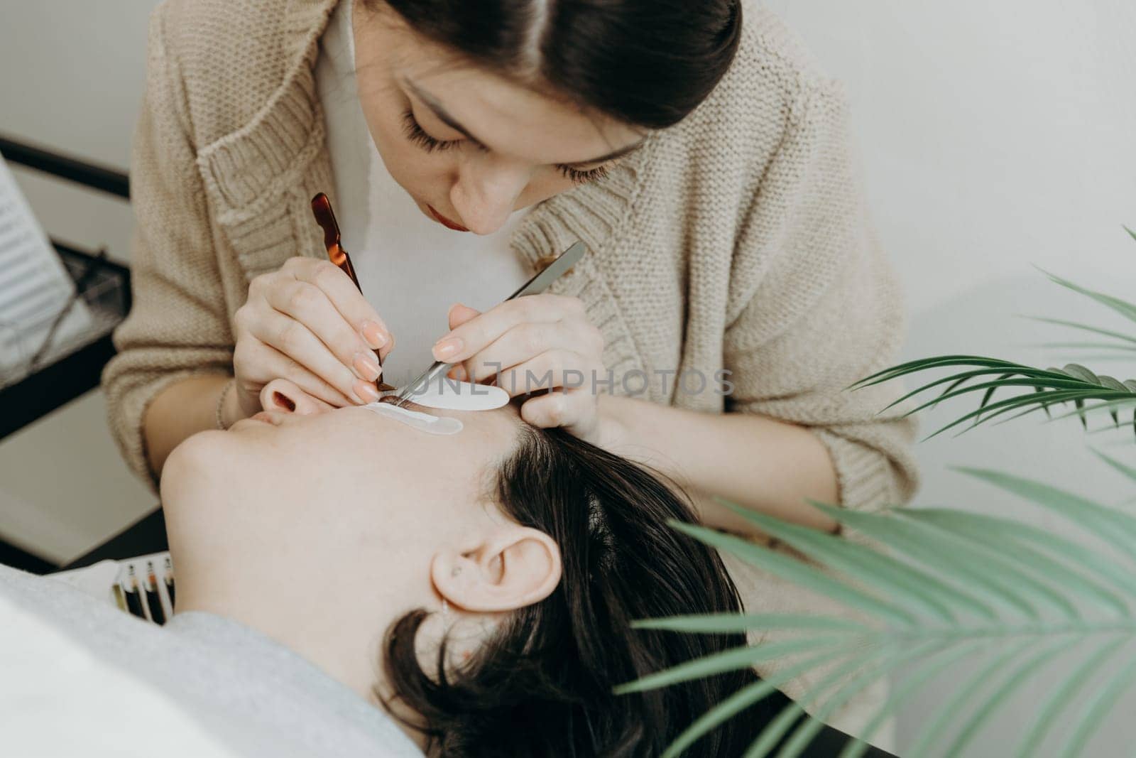 Portrait of one young beautiful Caucasian brunette girl, a cosmetologist glues artificial eyelashes with two tweezers to the left of a female client lying on a cosmetology table in the office on a spring day, side view, close-up. Concept of eyelash extensions, beauty industry.