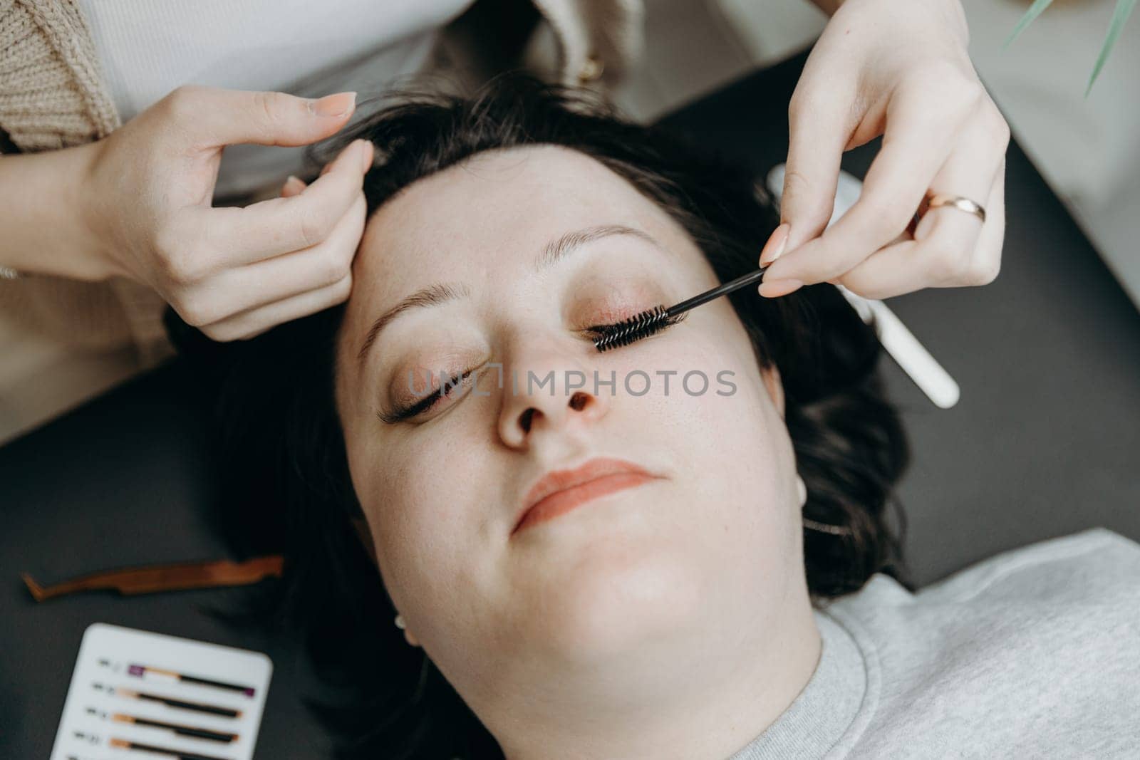 Portrait of one young unrecognizable Caucasian cosmetologist girl who combs artificial eyelashes with a brush on the left of a female client lying on the cosmetology table in the office on a spring day, top side close-up view. Concept of eyelash extensions, beauty industry.