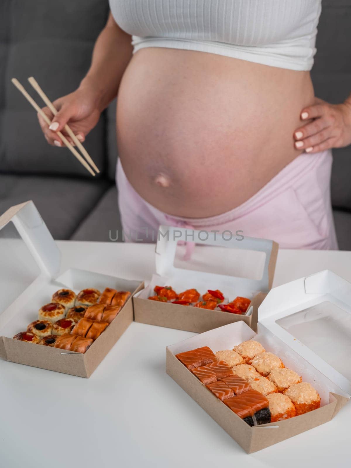 A pregnant woman holds chopsticks and stands at the table with rolls. Close-up of the belly. by mrwed54