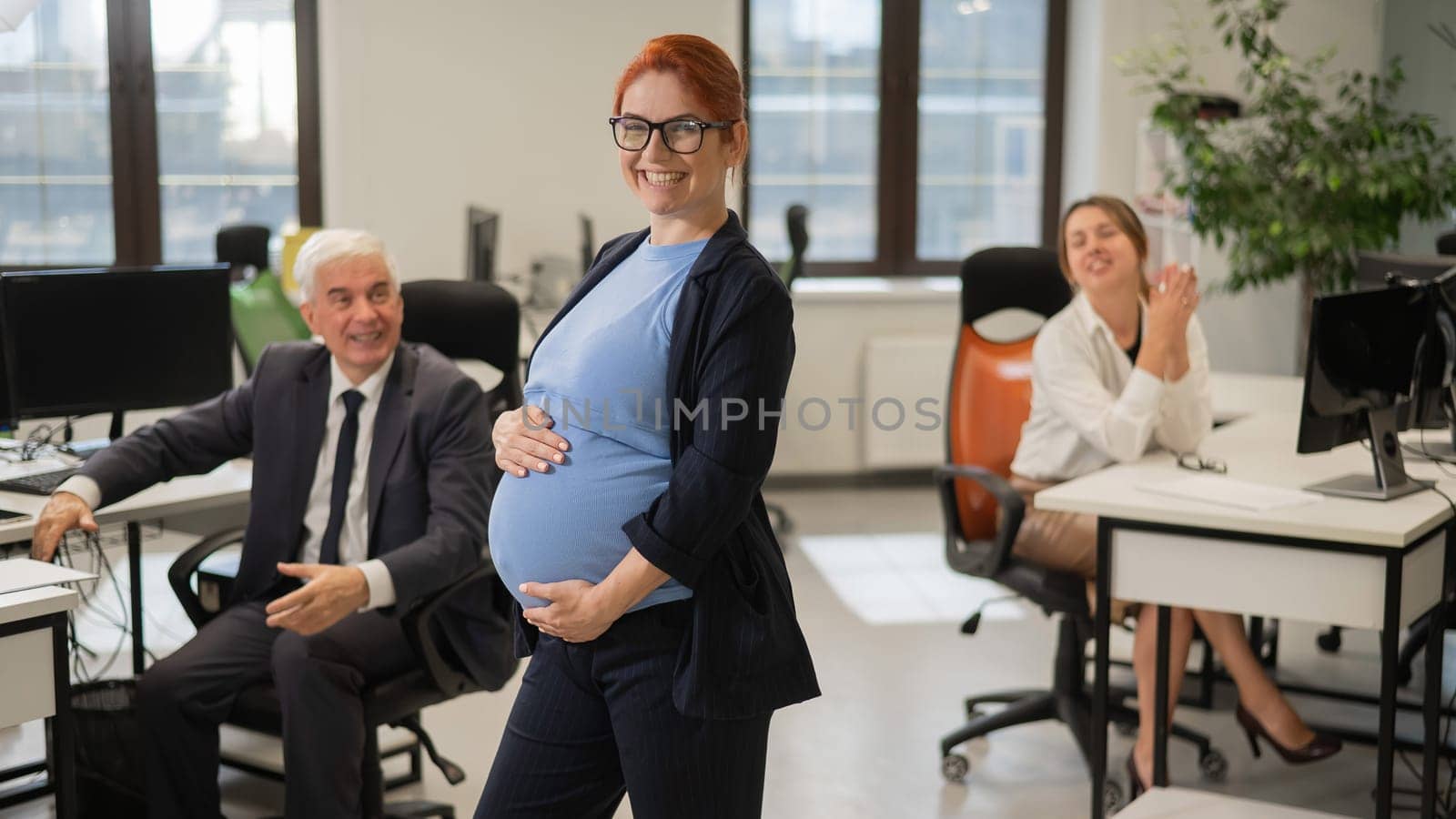 A happy pregnant woman stands in the middle of the office next to a Caucasian woman and an elderly man working at computers. by mrwed54