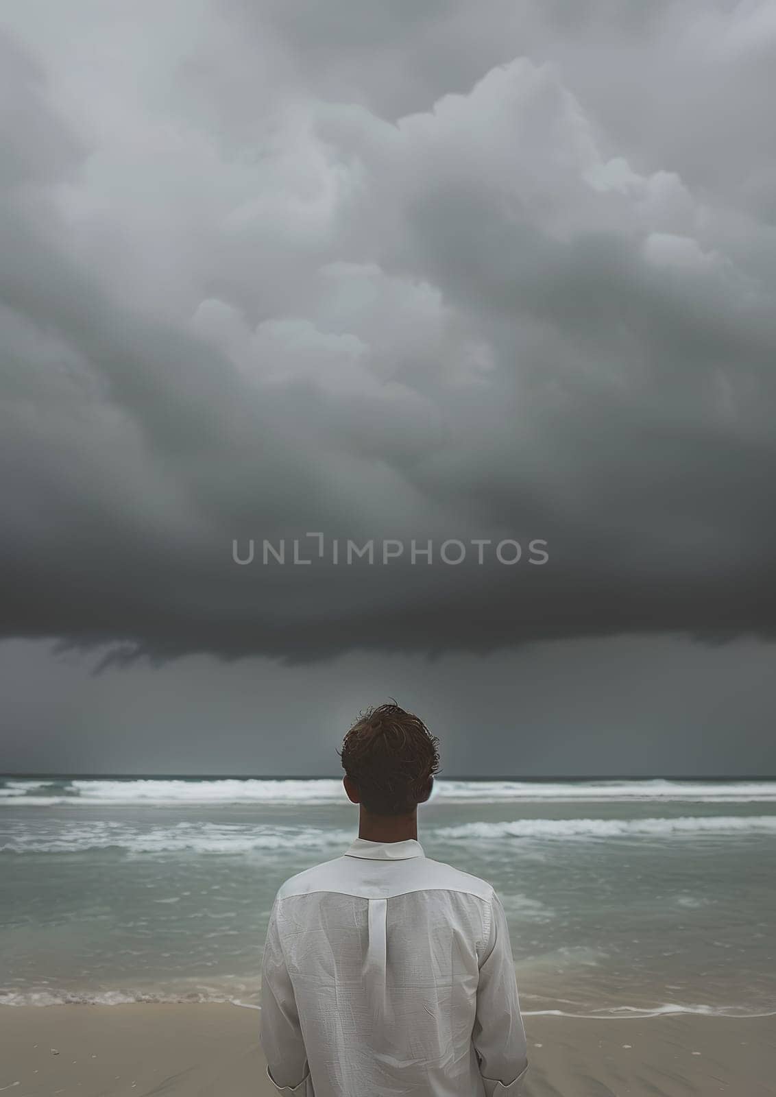 Man stands on coastal beach, gazing at ocean under cloudy sky by Nadtochiy