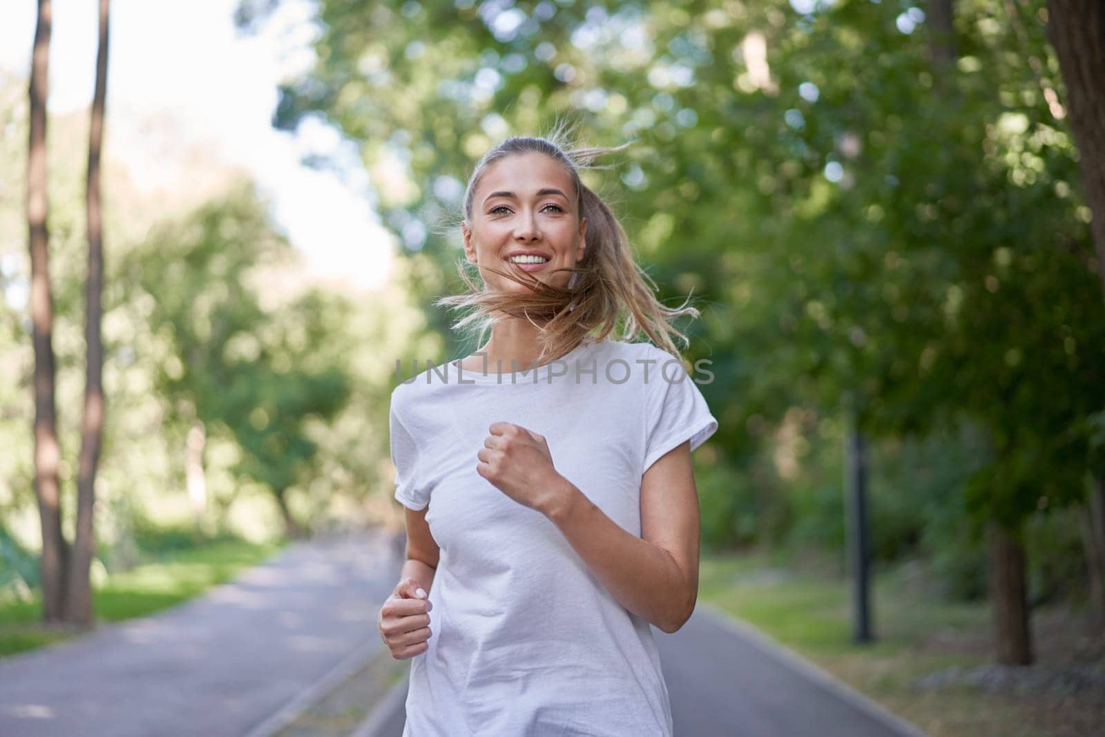 Portrait of smiling young sportswoman in white t-shirt jogging in park. Confident girl runner, jogger in sportswear running on road and looking at camera. Embracing a healthy lifestyle concept.