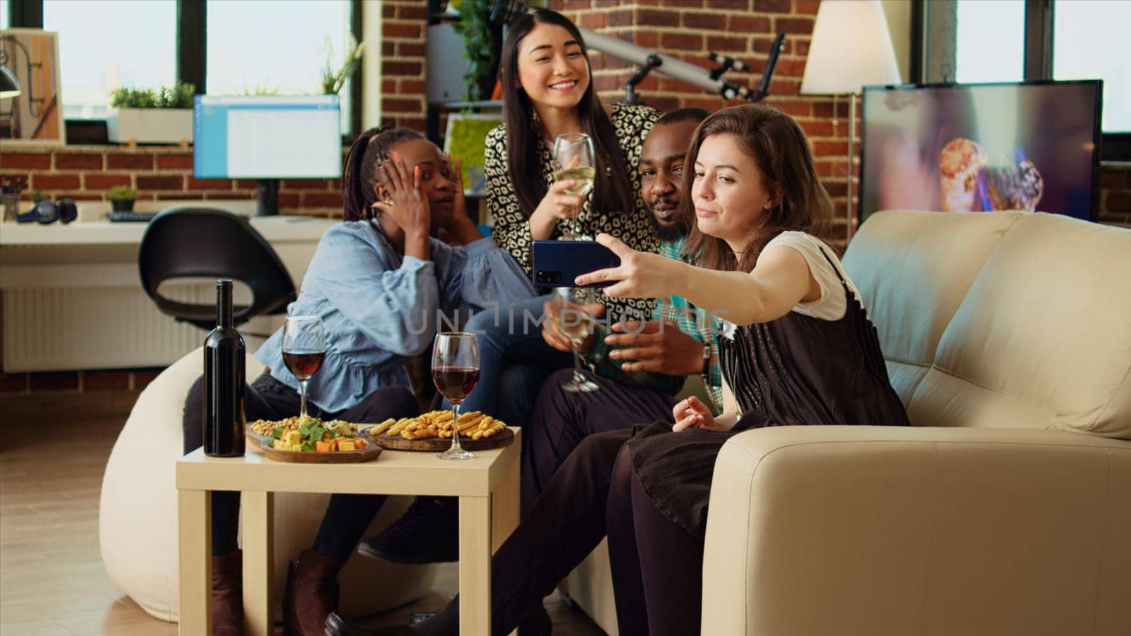 Multiethnic friends taking group pictures with cellphone at apartment gathering. Happy people in living room posing for photo, making funny faces and drinking alcoholic beverages