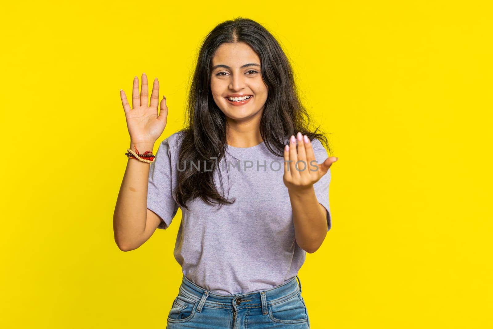 Young Indian woman smiling friendly at camera, waving hands gesturing hello greeting or goodbye welcoming with invitation hospitable expression. Arabian Hindu girl isolated on yellow studio background