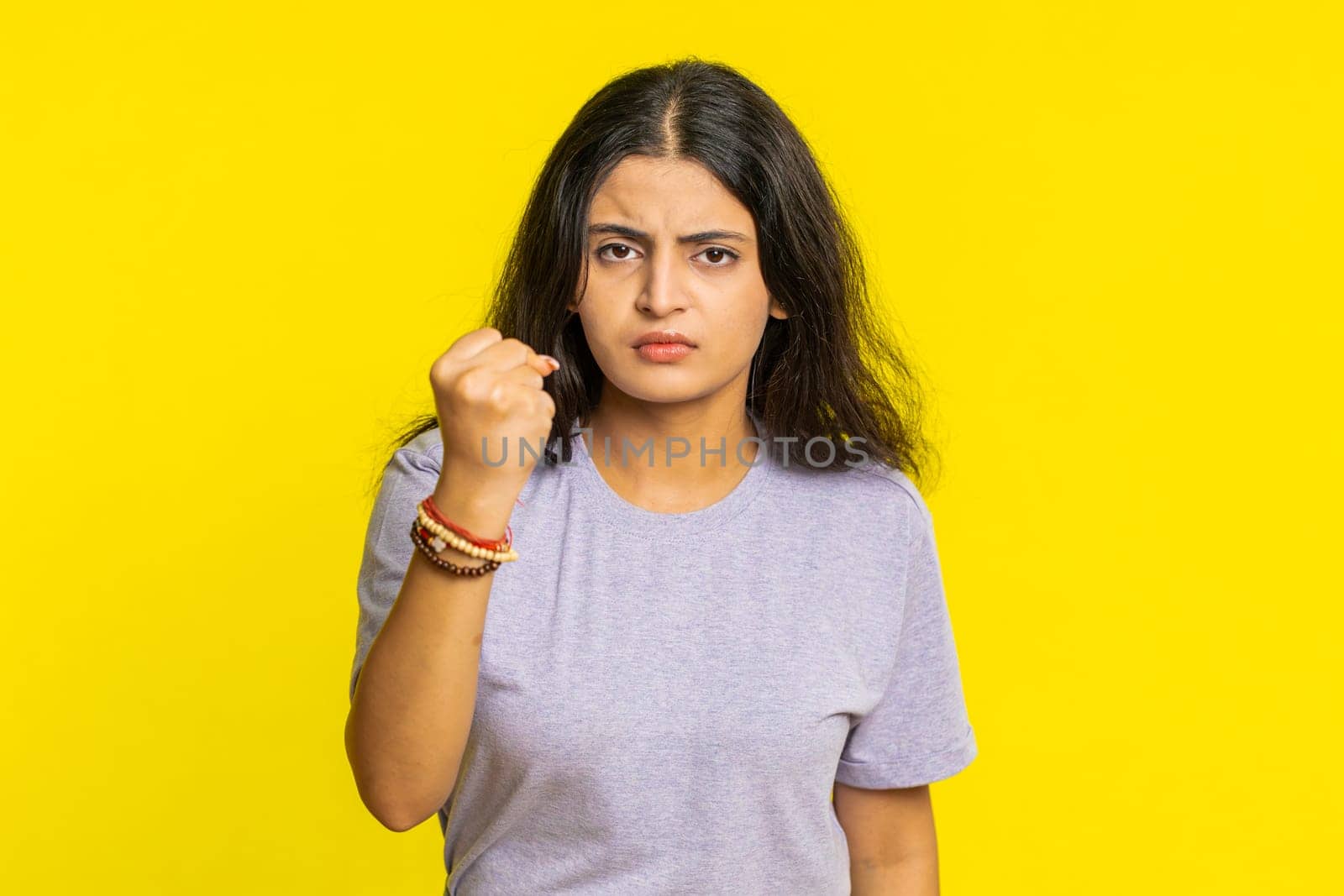 Aggressive angry Indian young woman trying to fight at camera, shaking fist, boxing with expression, punishment, threaten, bullying, abuse, mad fury. Arabian girl isolated on yellow studio background