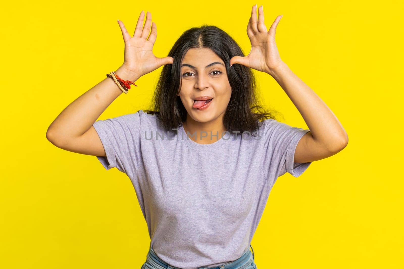 Playful Indian young woman showing tongue making faces at camera, fooling around, joking, aping with silly face, teasing, bullying, abuse. Cheerful funny Arabian girl isolated on yellow background