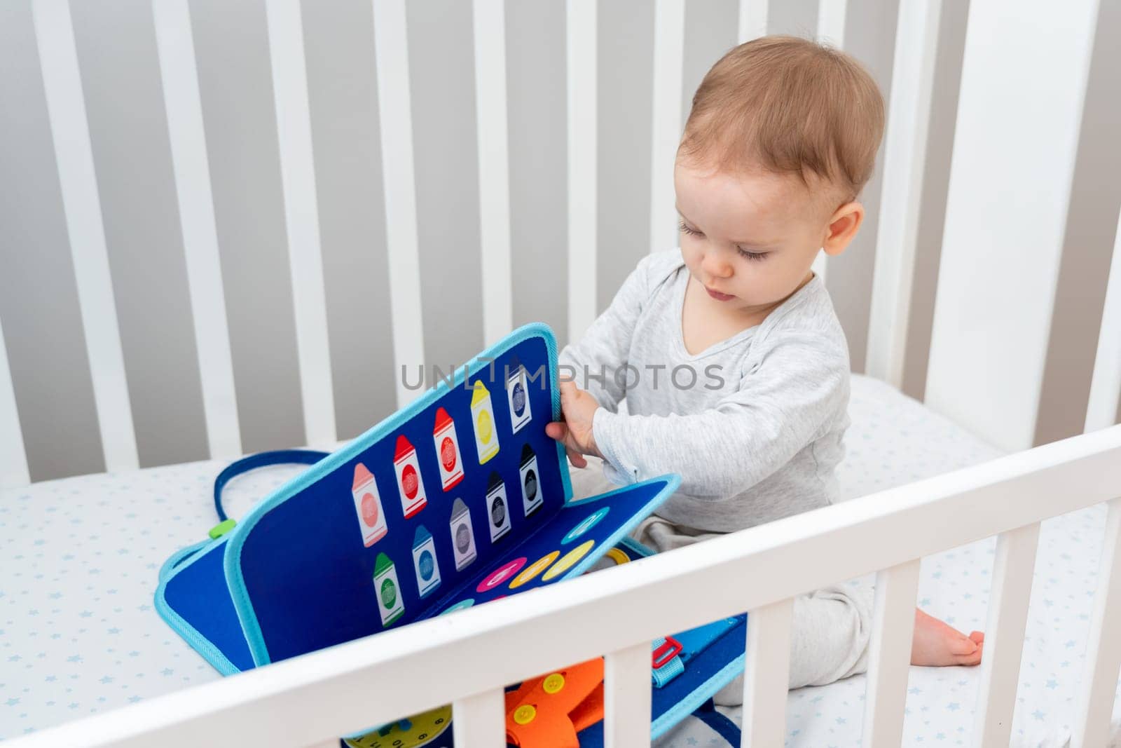 One year old baby playing with montessori busy book sitting in crib