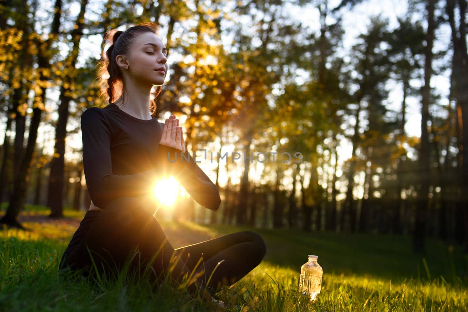 A young beautiful woman in a tight-fitting tracksuit sits on the grass in the woods in lotus pose and meditates