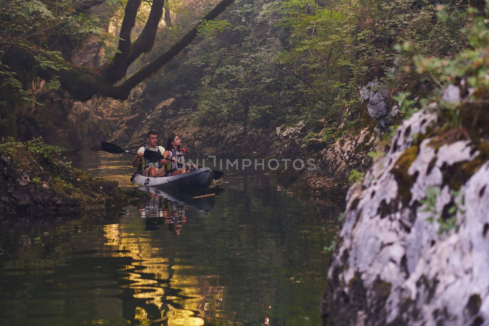 A young couple enjoying an idyllic kayak ride in the middle of a beautiful river surrounded by forest greenery.