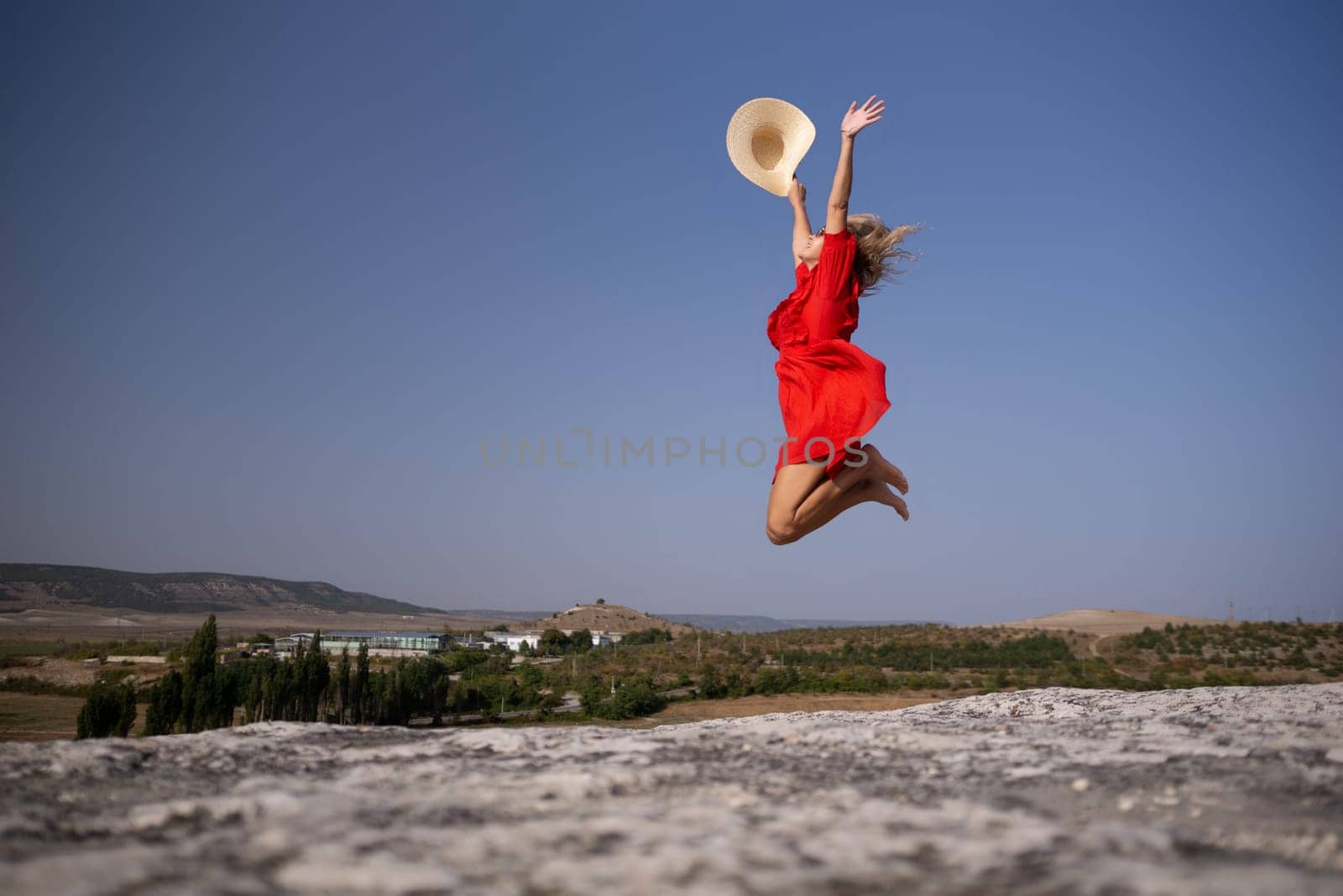 A woman in a red dress is jumping in the air with a straw hat on. The sky is clear and the sun is shining brightly. The woman's expression is joyful and carefree