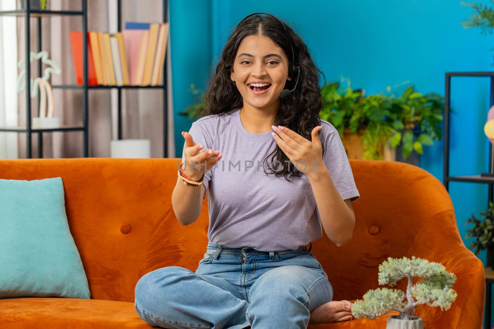 Indian woman wearing headset, freelance worker, call center or support service operator helpline. Arabian girl having talk with client or colleague communication support waving hello alone at home