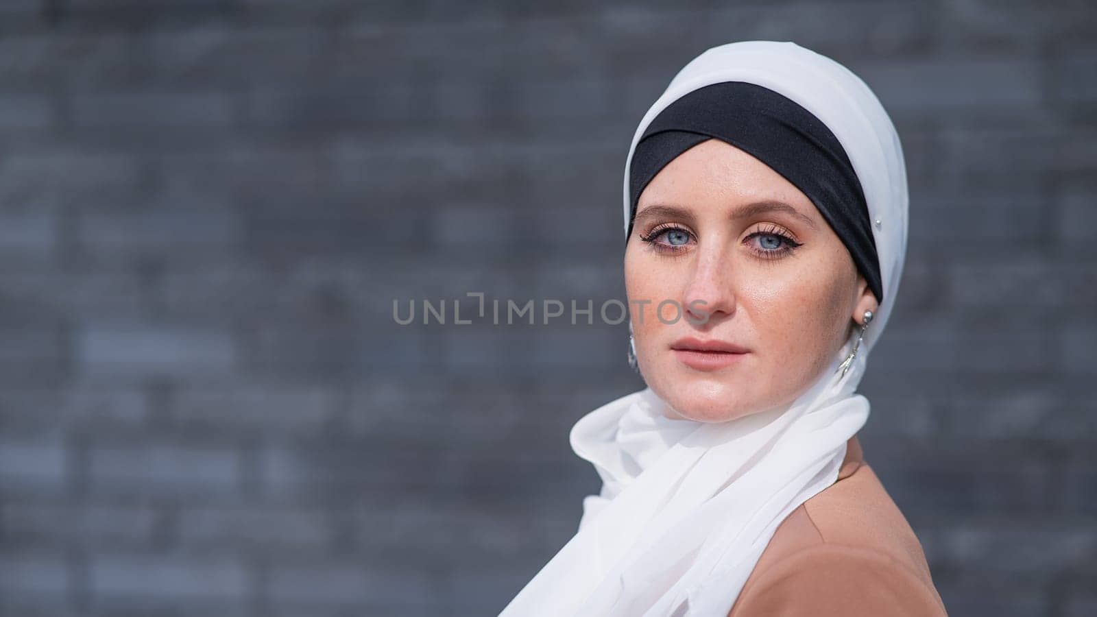 Portrait of a young blue-eyed woman in a hijab against a gray brick wall