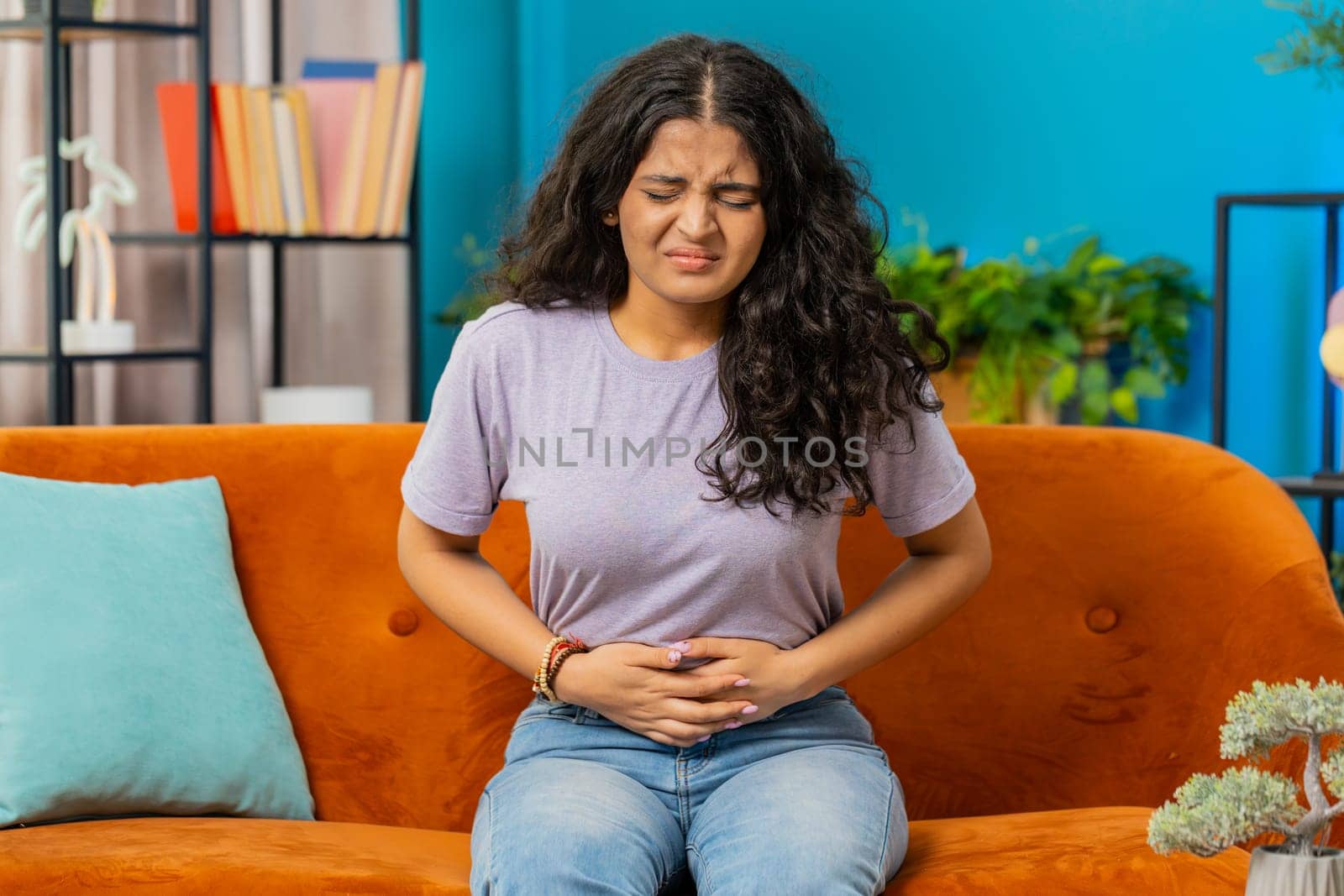 Sick ill Indian woman suffering from period cramps, painful stomach ache on sofa at home room. Girl holding belly, feeling abdominal menstrual pain. Abdominal pain, gastritis, diarrhea, indigestion