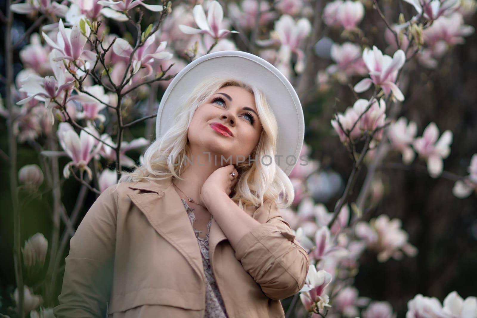 Magnolia flowers woman. A blonde woman wearing a white hat stands in front of a tree full of pink flowers. She is wearing a brown jacket and a necklace