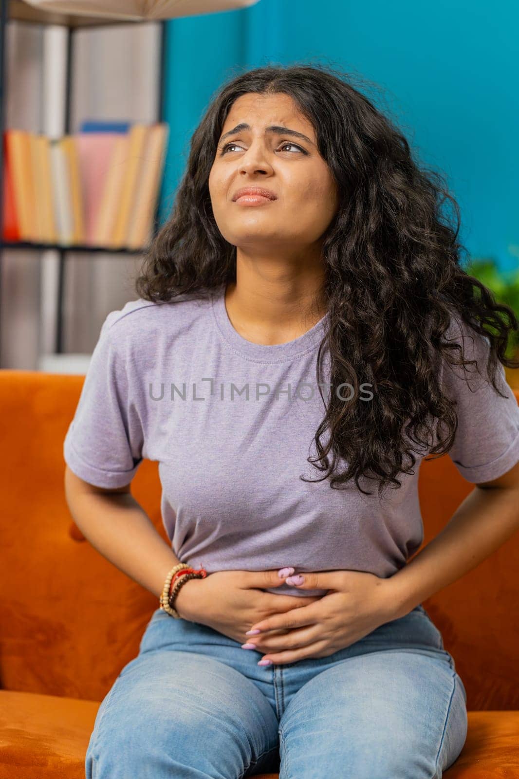 Sick ill Indian woman suffering from period cramps, painful stomach ache on sofa at home room. Girl holding belly, feeling abdominal menstrual pain. Abdominal pain, gastritis, diarrhea, indigestion