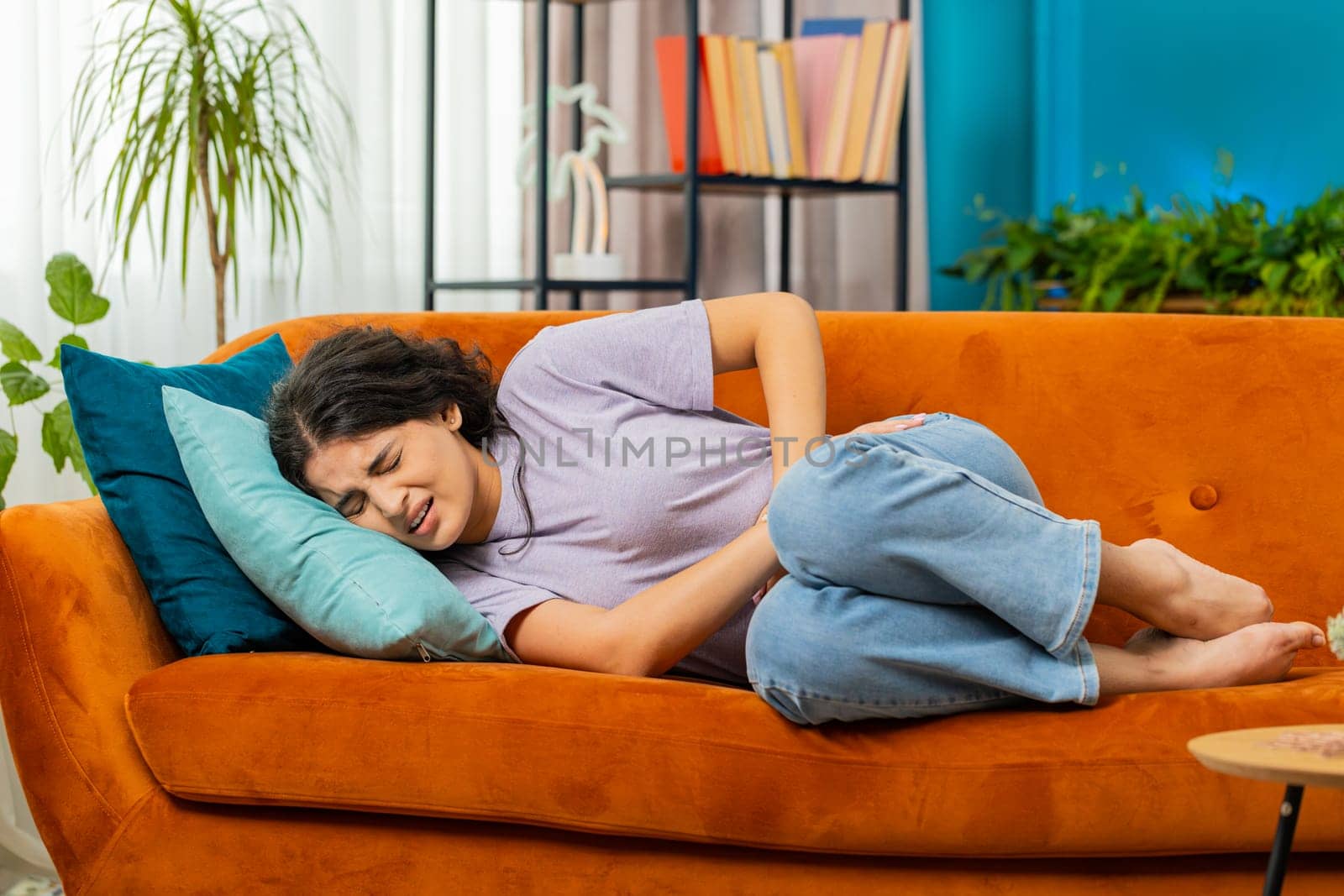 Sick ill Indian woman suffering from period cramps, painful stomach ache on sofa at home room. Girl holding belly, feeling abdominal menstrual pain. Abdominal pain, gastritis, diarrhea, indigestion