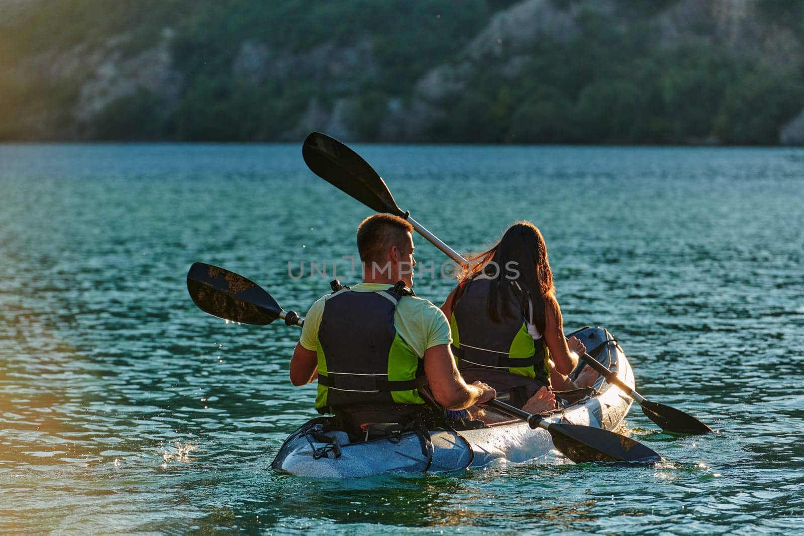 A young couple enjoying an idyllic kayak ride in the middle of a beautiful river surrounded by forest greenery in sunset time.