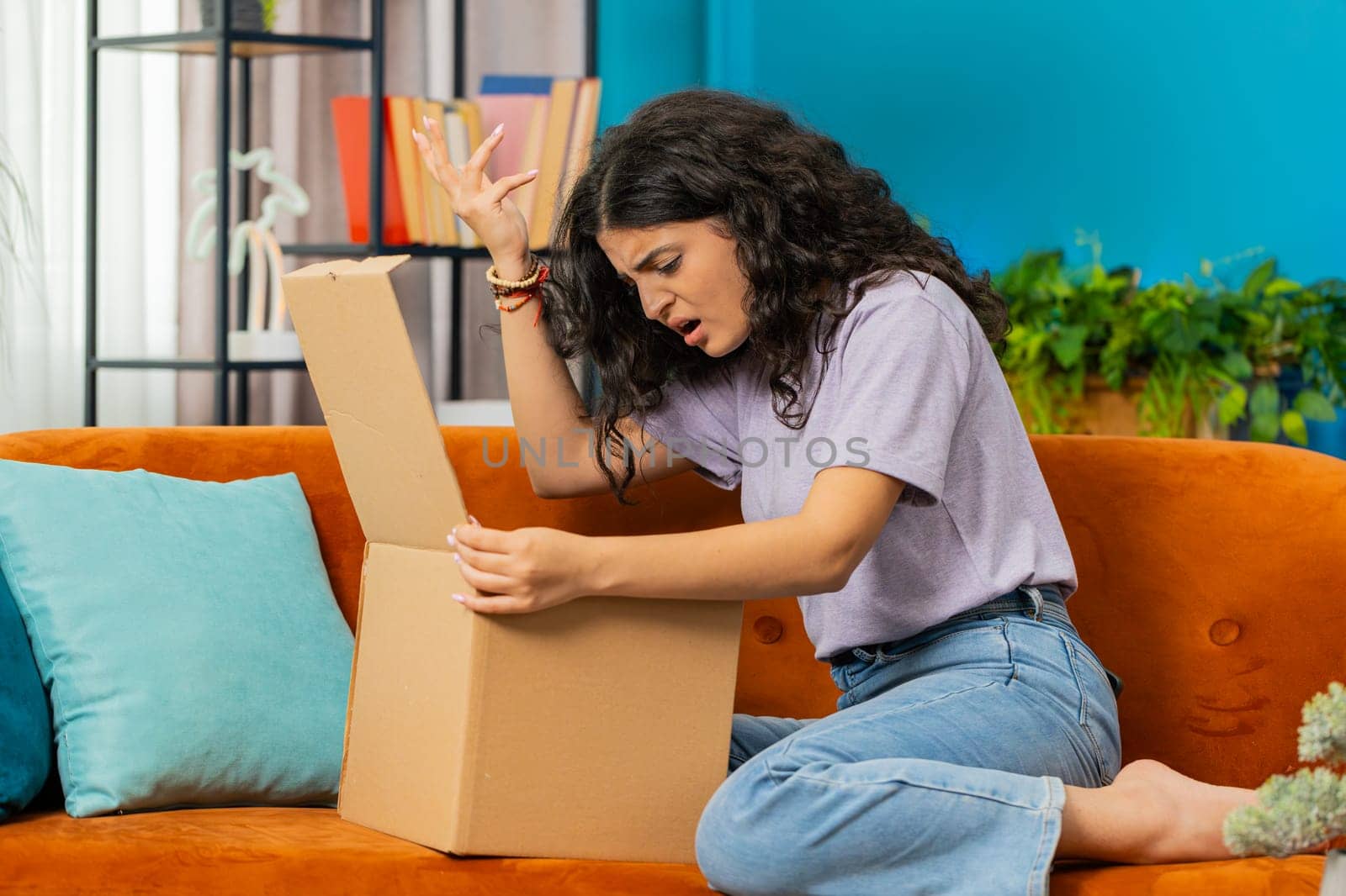 Angry dissatisfied shopper Indian woman unpacking parcel feeling upset and confused with wrong mistake delivery from an online store, bad quality broken purchase at home. Girl indoors in room on couch