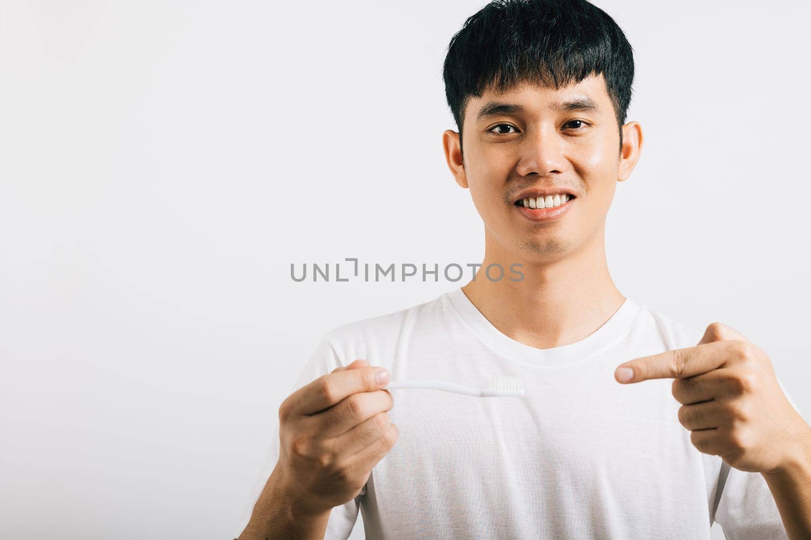 Smiling Asian teen brushes teeth in the morning, holding a toothbrush and pointing to it in a studio shot isolated on white background. Dental health concept with positive expression.
