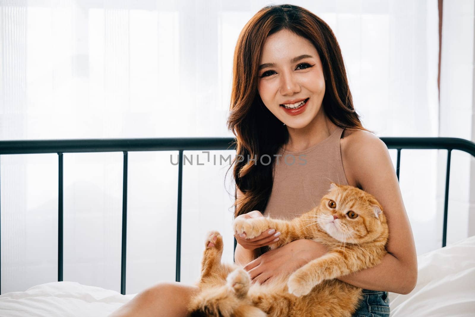 In the bedroom, a young woman smiles with delight while holding her cute Scottish Fold cat, exemplifying the affectionate bond between owner and pet, a portrait of happiness. Pet love by Sorapop