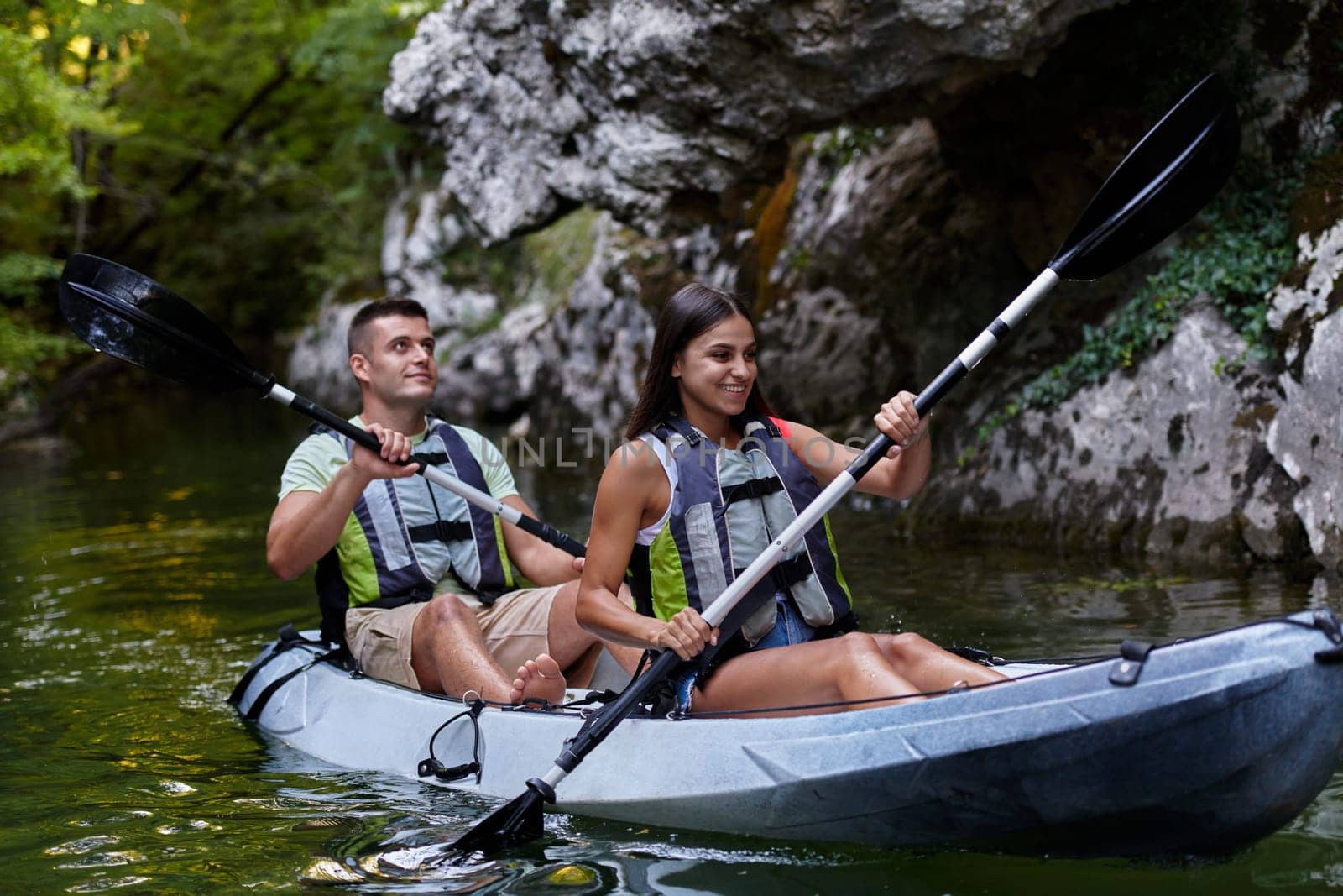 A young couple enjoying an idyllic kayak ride in the middle of a beautiful river surrounded by forest greenery.